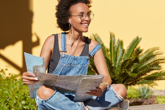 Mujer afroamericana satisfecha negra usa el mapa de destino para buscar lugares interesantes, busca turismo en un lugar desconocido, disfruta de la transmisión de radio en auriculares, posa al aire libre