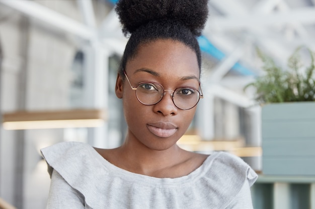 Mujer afroamericana seria de piel bastante oscura con labios gruesos, usa gafas redondas, posa en interiores, se sienta contra un espacioso espacio de oficina