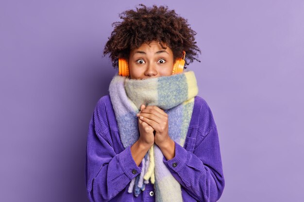 Mujer afroamericana siente mucho frío durante el clima helado usa chaqueta púrpura y bufanda cálida alrededor del cuello en la calle durante el invierno escucha música a través de auriculares inalámbricos mantiene las manos juntas