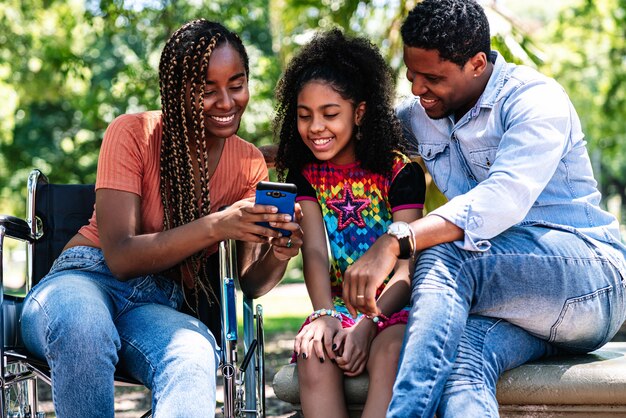 Una mujer afroamericana en silla de ruedas disfrutando de un día en el parque con su familia mientras usa un teléfono móvil juntos.