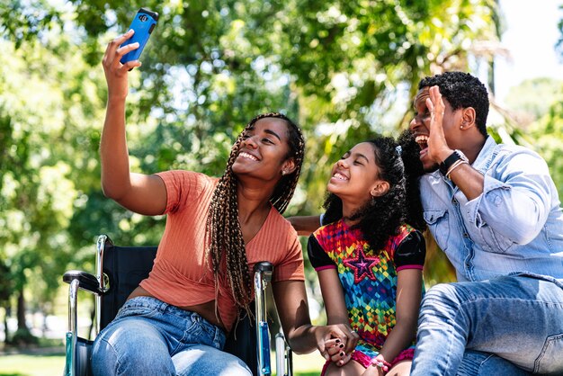 Una mujer afroamericana en silla de ruedas tomando un selfie con su familia con un teléfono móvil mientras disfruta de un día en el parque.