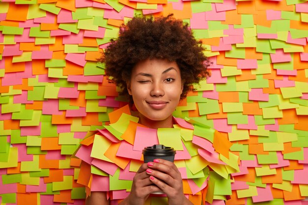 Mujer afroamericana soñolienta guiña un ojo, sostiene una taza de café desechable, trabaja durante muchas horas, trata de estar fresca, tiene el cabello rizado natural, pega la cabeza a través del fondo de papel, notas adhesivas alrededor