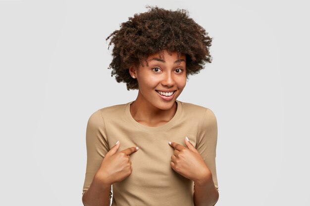 La mujer afroamericana sonriente positiva se indica a sí misma, expresa sorpresa, no puede creer que un amigo se enamore de ella, mira con incredulidad, se para contra la pared blanca