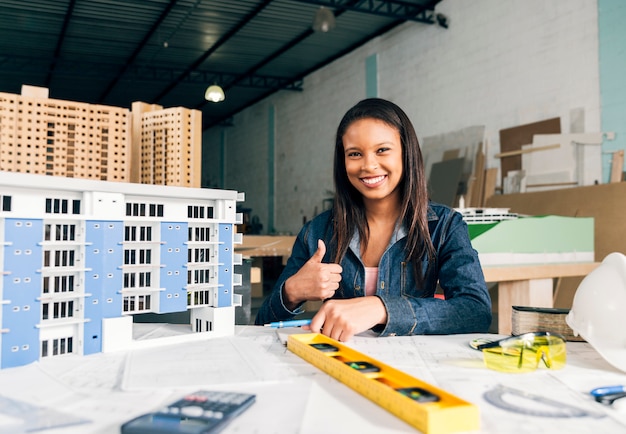 Mujer afroamericana sonriente que muestra el pulgar hacia arriba cerca del modelo de edificio