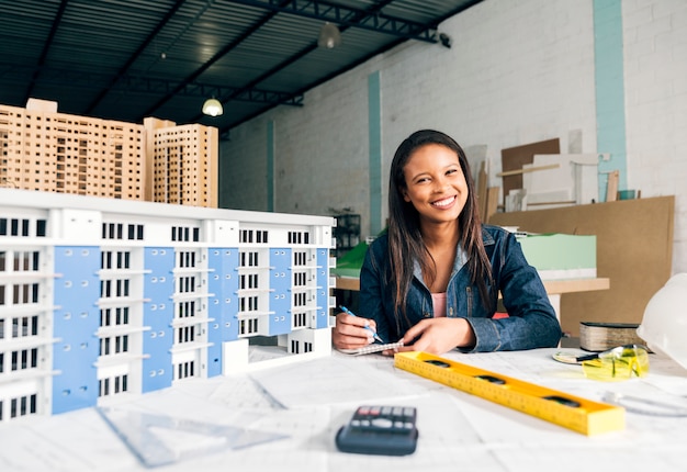 Mujer afroamericana sonriente tomando notas cerca del modelo de edificio
