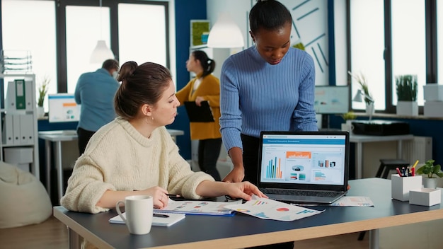 Mujer afroamericana sosteniendo una computadora portátil preguntando a un colega mirando el portapapeles con gráficos para obtener consejos sobre datos financieros en una oficina ocupada. Dos colegas que analizan la cuota de mercado intercambian ideas.