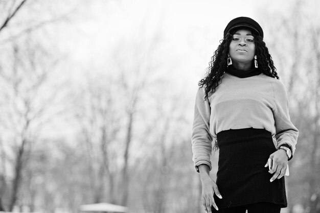 Mujer afroamericana con suéter marrón de falda negra y gorra posada en el día de invierno contra un fondo nevado sosteniendo el teléfono a mano