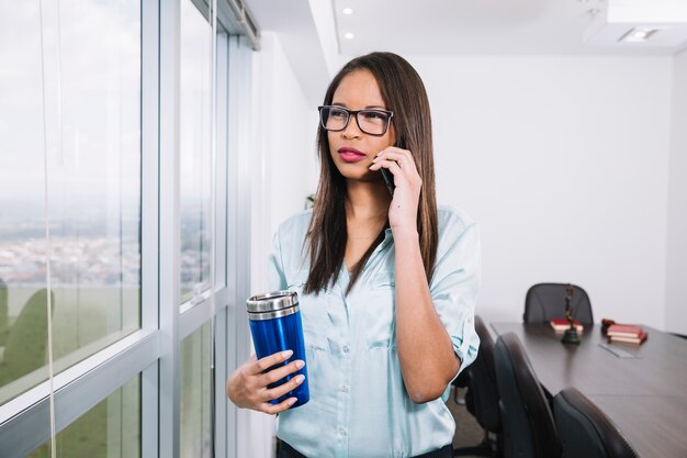 Mujer afroamericana con termo hablando en teléfono inteligente cerca de la ventana en la oficina