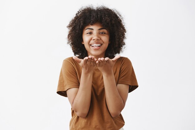 Mujer afroamericana tierna y romántica despreocupada que expresa afecto y amor mientras envía un beso de viento o mwah sonriendo con alegría y sosteniendo las palmas cerca de la boca sobre una pared gris