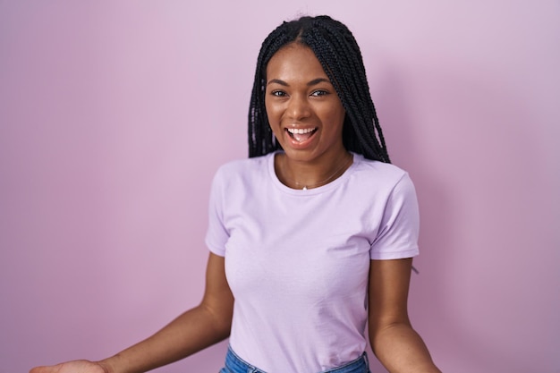 Mujer afroamericana con trenzas sobre fondo rosa sonriendo alegre con los brazos abiertos como bienvenida amistosa, saludos positivos y confiados