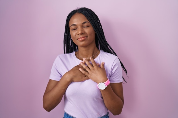 Mujer afroamericana con trenzas sobre fondo rosa sonriendo con las manos en el pecho con los ojos cerrados y un gesto agradecido en la cara. concepto de salud