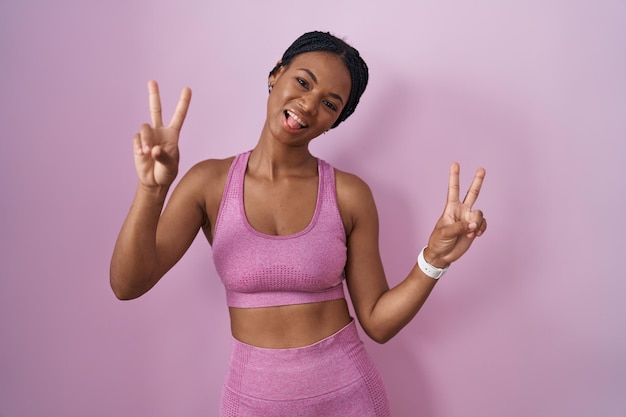 Mujer afroamericana con trenzas usando ropa deportiva sobre fondo rosa sonriendo con la lengua afuera mostrando los dedos de ambas manos haciendo el signo de la victoria. número dos.