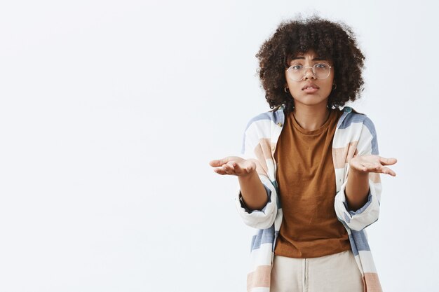 Mujer afroamericana triste y sombría desesperada con gafas transparentes con el pelo rizado levantando las palmas cerca del pecho en un gesto confuso y desorientado, no puedo entender por qué