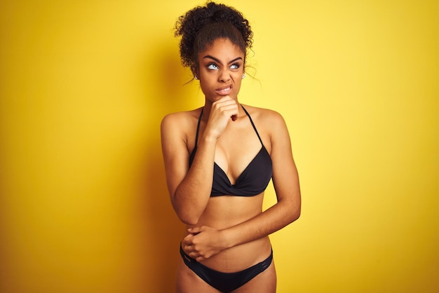 Foto gratuita mujer afroamericana de vacaciones con bikini de pie sobre un fondo amarillo aislado pensando preocupada por una pregunta preocupada y nerviosa con la mano en la barbilla