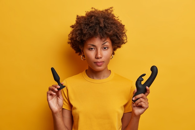 Foto gratuita la mujer afroamericana vacilante sostiene un tapón anal para calentar antes del juego con penetración, un vibrador en forma de conejo para la estimulación de la vagina, vestida con una camiseta amarilla. mujer joven, con, juguetes sexuales, interior