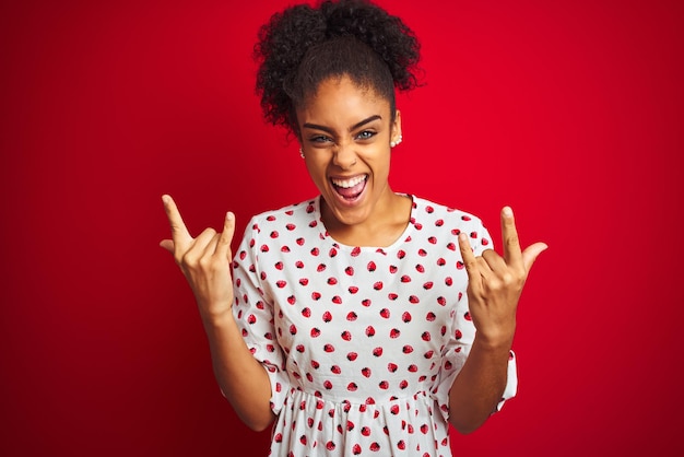 Mujer afroamericana con vestido blanco de moda de pie sobre fondo rojo aislado gritando con expresión loca haciendo símbolo de rock con las manos arriba Estrella de la música Concepto pesado