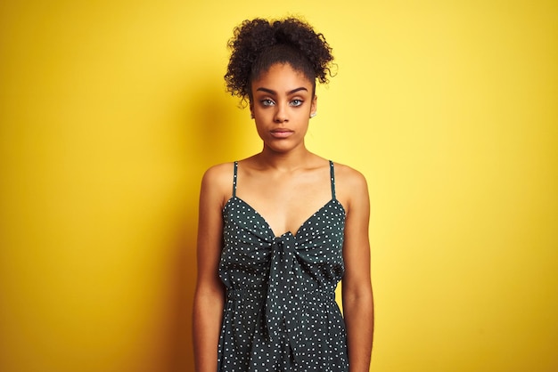 Mujer afroamericana con vestido verde casual de verano sobre fondo amarillo aislado Relajada con expresión seria en la cara Mirada simple y natural a la cámara