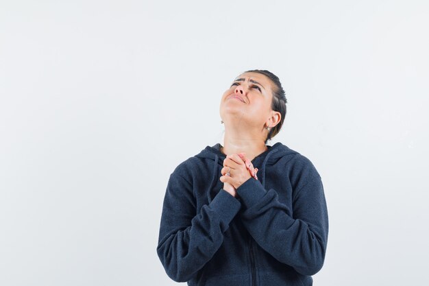 Mujer agarrando las manos en gesto de oración en sudadera con capucha y mirando esperanzado, vista frontal.