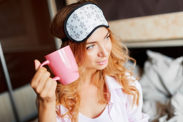 Mujer agraciada con sincera sonrisa posando en máscara de ojos en su habitación y beber té caliente.