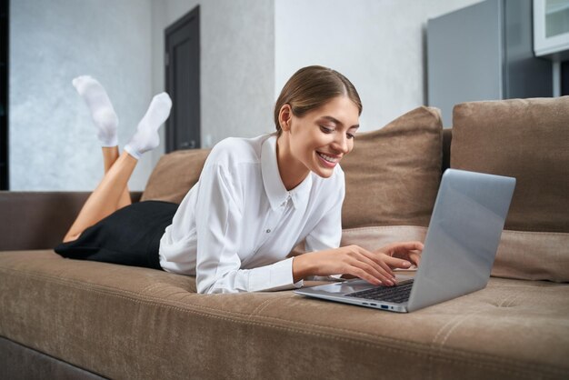 Mujer agradable acostada en el sofá y escribiendo en la computadora portátil