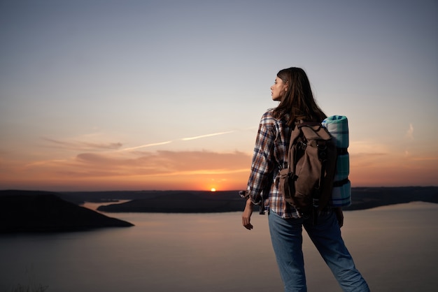 Mujer agradable con mochila disfrutando del atardecer desde la colina