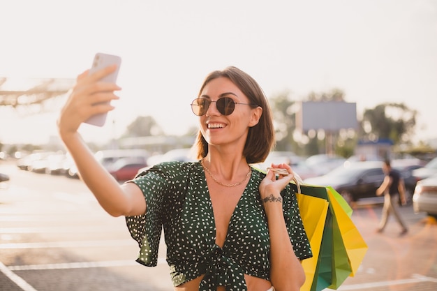 Mujer al atardecer con coloridas bolsas de compras y estacionamiento por centro comercial feliz con teléfono móvil tomar foto selfie