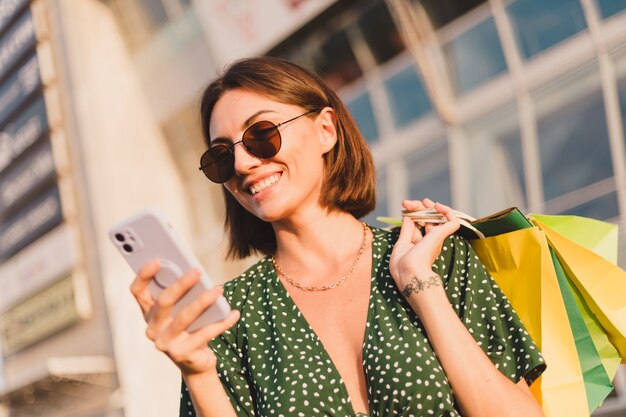 Mujer al atardecer con coloridas bolsas de compras y estacionamiento por centro comercial feliz con teléfono móvil