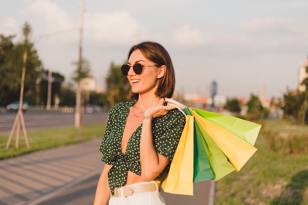 Mujer al atardecer con coloridas bolsas de compras en el parque de la calle de la ciudad después del día de compras feliz