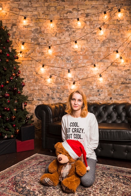 Mujer al lado de árbol de navidad con peluche que lleva gorro de navidad