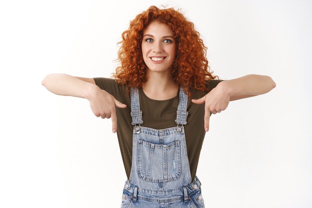 Mujer alegre, amistosa y optimista con un corte de pelo rizado de jengibre que muestra un anuncio en la parte inferior, sonriendo y mirando al frente feliz, apuntando hacia abajo, da consejos, usa el código de promoción, recomienda ver videos de tendencias