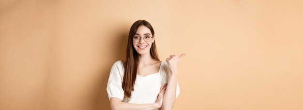 Mujer alegre en anteojos señalando con el dedo a la izquierda en el logo sonriendo feliz de pie sobre fondo beige