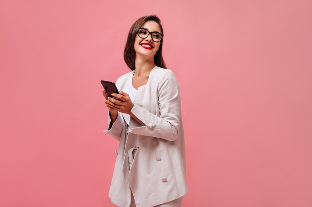 Mujer alegre en anteojos y traje beige sostiene smartphone sobre fondo rosa. Mujer de negocios sonriente en traje elegante posando en cámara.