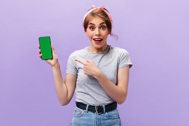 Mujer alegre apunta al teléfono sobre fondo púrpura. Encantadora chica con peinado fresco con pañuelo en traje de verano mirando a cámara.