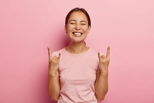 Mujer alegre de aspecto agradable hace letrero de rock, gesticula con ambas manos, se siente rebelde, muestra heavy metal, se vuelve loca, escucha música alta en casa, se viste de manera informal. Chica asiática se divierte en la fiesta