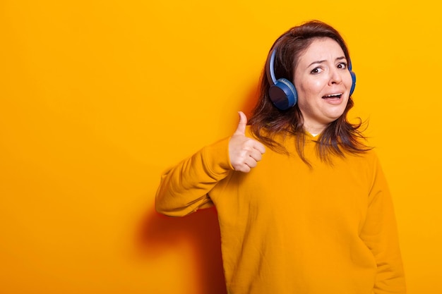 Mujer alegre con auriculares para escuchar música y mostrando el símbolo bien con la mano en la cámara. Adulto positivo que se siente feliz y escucha canciones y sonidos en los auriculares, divirtiéndose