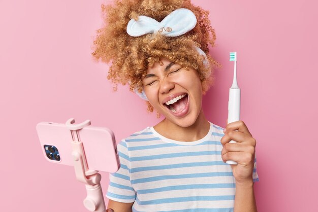 Una mujer alegre y bastante atractiva se siente muy feliz graba un video para el blog y da recomendaciones sobre cómo usar el cepillo de dientes eléctrico usa una diadema y una camiseta casual a rayas aislada sobre un fondo rosa