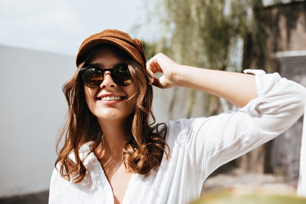 Mujer alegre en blusa blanca, ligera con cuello en V con una sonrisa mirando a la cámara. Chica con gorra y gafas posando contra el espacio del edificio y los árboles.