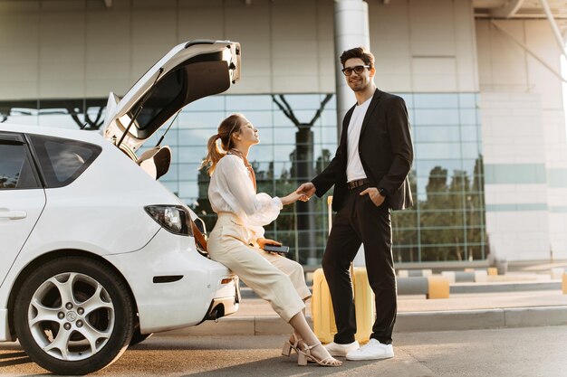 Mujer alegre en blusa blanca pantalones beige se sienta en el auto y sostiene la mano del novio Hombre morena en traje negro y anteojos sonríe y posa cerca del aeropuerto