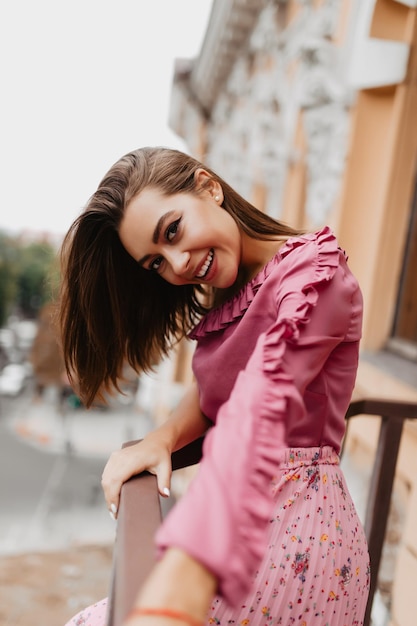 Una mujer alegre con una blusa suave y tierna mira a la cámara con coquetería Retrato de una mujer europea con estilo que se extiende hacia la cámara en el balcón