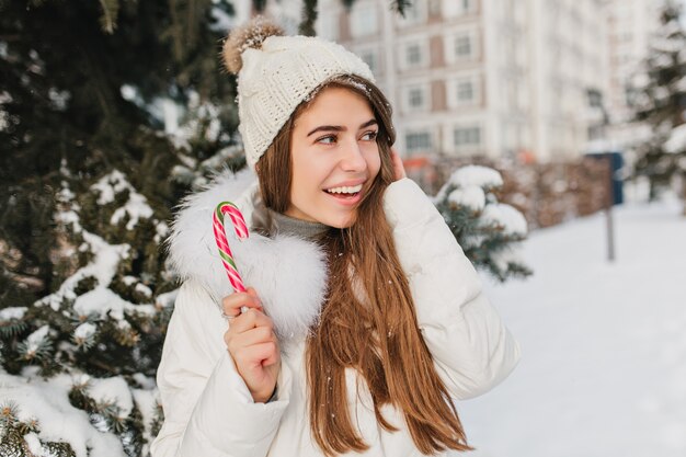 Mujer alegre con cabello largo brillante sosteniendo bastón de caramelo y mirando a otro lado. Foto al aire libre de guapa mujer rubia con piruleta disfrutando de las vacaciones de invierno.