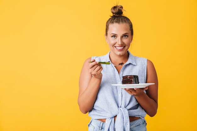 Mujer alegre con camisa mirando alegremente a la cámara sosteniendo un plato con una rebanada de pastel de chocolate en las manos sobre un fondo amarillo aislado