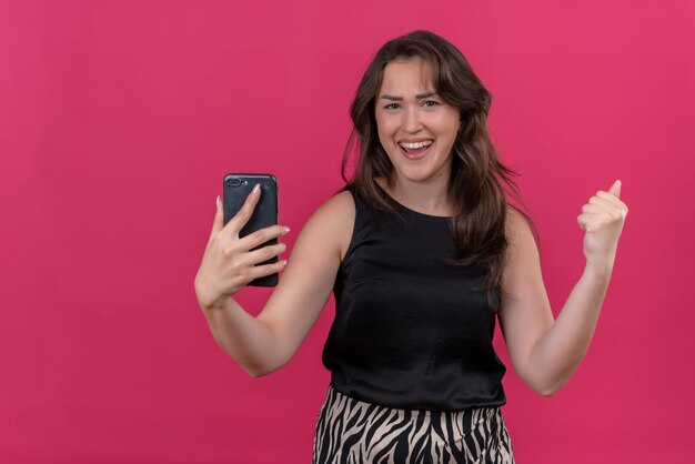 Mujer alegre con camiseta negra escucha música desde el teléfono y baila en la pared rosa