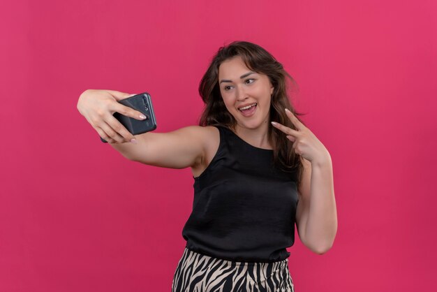 Mujer alegre con camiseta negra tomar un selfie y mostrar gesto viva en la pared rosa
