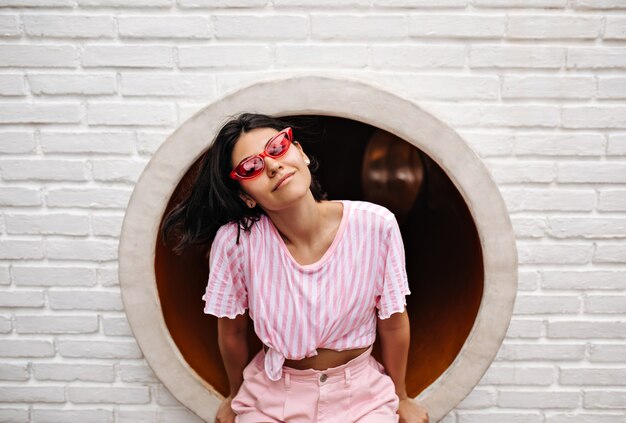 Mujer alegre en camiseta sentada en la pared de ladrillos. Tiro al aire libre de mujer encantadora en gafas de sol de moda.