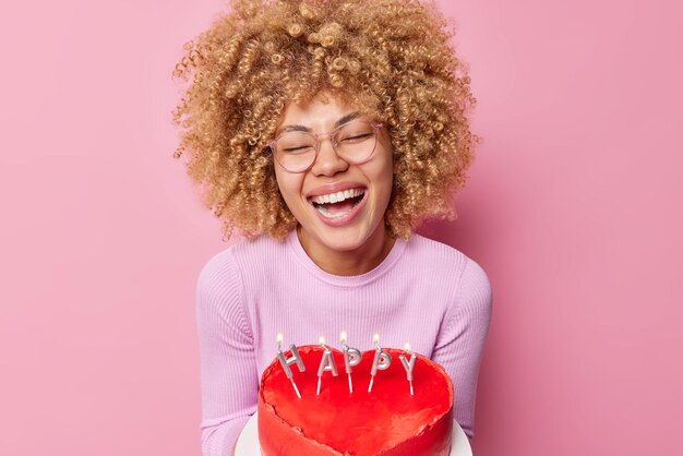 Una mujer alegre celebra el aniversario de salir con su novio sostiene un pastel en forma de corazón con velas encendidas y se ríe positivamente usa anteojos transparentes y un puente aislado en el fondo del estudio rosa