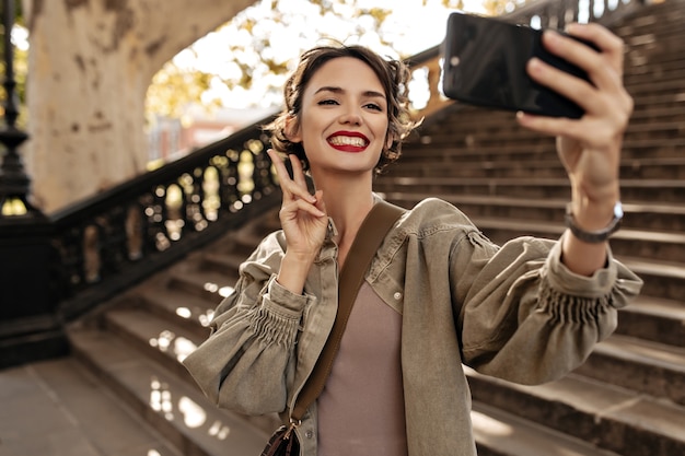 Mujer alegre en chaqueta de mezclilla verde oliva mostrando el signo de la paz y sonriendo afuera. Mujer de pelo corto con labios rojos haciendo selfie de escaleras.