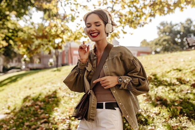 Mujer alegre en chaqueta verde oliva y jeans blancos sonriendo afuera. Mujer de pelo ondulado en auriculares con bolso escuchando música al aire libre.