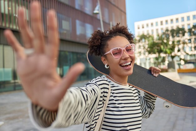 Mujer alegre despreocupada mantiene la palma hacia adelante cámara sonríe positivamente disfruta andar en patineta usa gafas de sol rosas y un puente de rayas casuales camina al aire libre