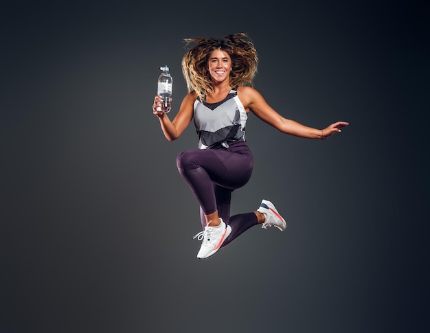 Una mujer alegre y emocionada salta al estudio mientras sostiene la botella de agua sobre un fondo gris.