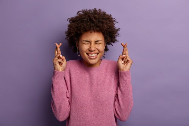 Foto gratuita una mujer alegre, emotiva y complacida ora a dios por buena suerte, cruza los dedos, sonríe ampliamente, muestra los dientes blancos, usa un jersey rosado